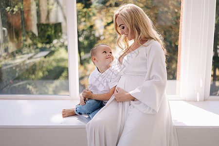 美丽的孕妇和她的儿子 男孩拥抱他的妈妈并且高兴 男孩在等姐姐 穿着优雅的白色连衣裙的女士在工作室里向摄影师摆姿势 白色薄纱背景成图片