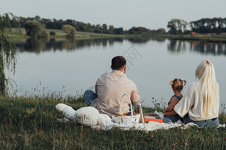 有野餐时间的年轻家庭的背影 爸爸妈妈和他们的小女儿坐在城外的湖边图片
