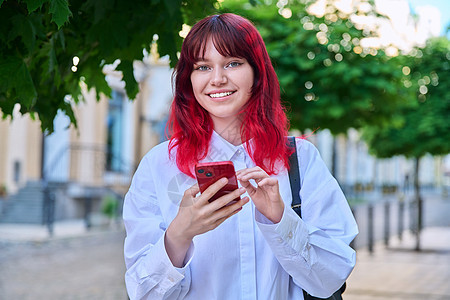 女性青少年的户外肖像画 手握智能手机在城内观看照相机青年女士女孩微笑发型时尚冒充学生技术红发图片