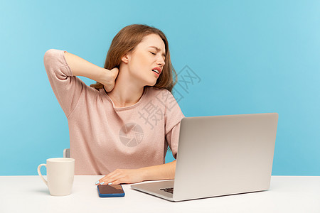 情感年轻女性在蓝背景的笔记本电脑上工作人士疾病鬼脸紧张背痛疼痛脊柱脖子伤害肩膀图片