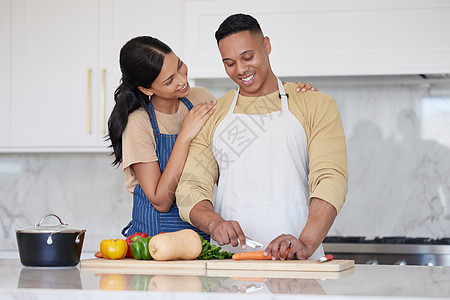 一对年轻的拉丁情侣在家里一起做一顿健康的晚餐 一个有爱心的男人切胡萝卜和其他蔬菜 准备饭菜 他的女朋友抱着他 在厨房里微笑图片