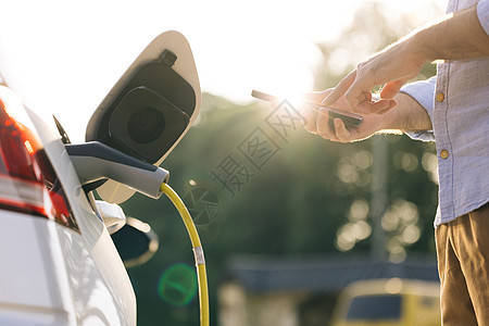 男性使用智能手机上的应用程序拔下电动汽车的电源线 商务人士在户外充电站为电动汽车充电图片