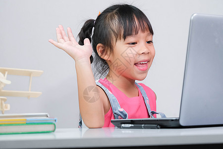 上学小女孩微笑的亚洲小女孩在线学习 与老师使用笔记本电脑进行视频通话 快乐的小女孩在家里用笔记本电脑在线学习时向导师挥手致意 在家上学电话背景