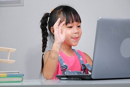 微笑的亚洲小女孩在线学习 与老师使用笔记本电脑进行视频通话 快乐的小女孩在家里用笔记本电脑在线学习时向导师挥手致意 在家上学女孩图片