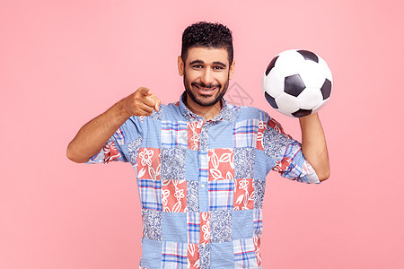 年轻的成年胡子男人带着露齿的微笑 穿着蓝色休闲风格的衬衫 指着你 拿着足球 用快乐的表情看着镜头中东高清图片素材
