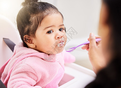 一名混血女婴在家中被妈妈用勺子喂食 可爱的蹒跚学步的小孩嘴里塞满了粥 坐在喂食椅上享用一些软食 营养促进健康发展图片