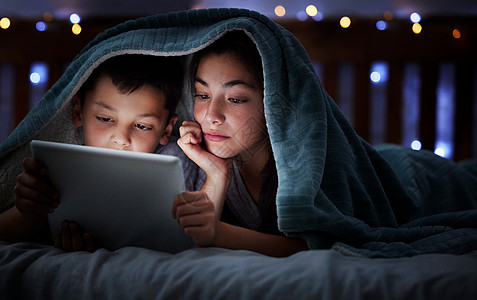 幸福的白人家庭有两个孩子 晚上在黑暗中躺在毯子下使用数字平板电脑 他们的脸被设备屏幕灯照亮 一家四口在睡前阅读网络故事或看电影青图片