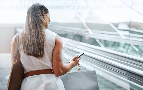 一名年轻女子在商场提着购物袋的自动扶梯上使用手机的后视图 女性一边享受购物疗法 一边与智能手机保持联系 使用应用程序查找销售和折图片