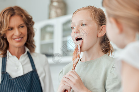 我必须品尝以确保它的美味 一个小女孩在家里和她的姐姐和祖母一起烘烤时舔着木勺图片