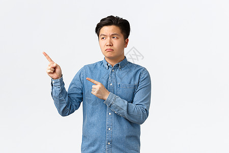 穿着蓝色衬衫的亚洲阴郁男人不高兴和失望 皱着眉头 心烦意乱 手指左上角 抱怨劣质产品 感到沮丧 站在白色背景背景图片