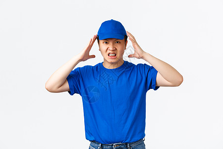 身穿蓝色制服的愤怒愤怒的亚洲送货员在头附近握手 疯狂和愤怒地做着鬼脸 快递员看起来很苦恼 厌倦了粗鲁的顾客 站在白色背景男人工作图片