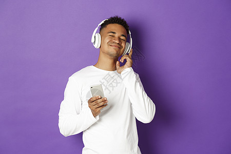 享受耳机听音乐 手持智能手机和微笑的欢笑 站在紫色背景上站立着 Y LAK购物男性学生网络成人情感多样性发型潮人员工背景图片