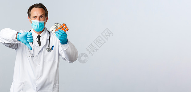 错误戴口罩Covid19 预防病毒 医护人员和疫苗接种概念 戴医用口罩和手套的严肃医生不高兴 展示大拇指和药物 劣质药丸 错误处方防护服隔背景