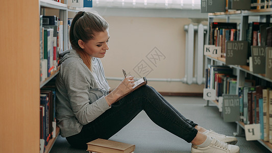 年轻漂亮的梦幻女学生坐在书架间轻盈图书馆的地板上 在抄本上写下作文 抬头看着天花板图片