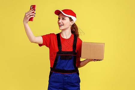 乐观的快递妇女站着拿着纸板盒 客户包裹和通过电话自拍图片