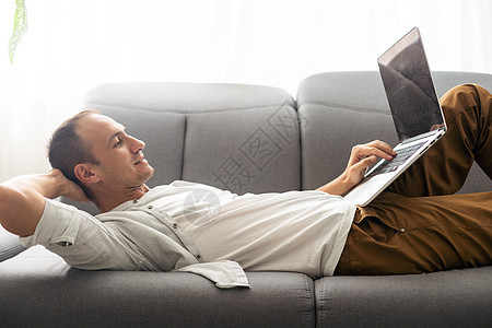 年轻有魅力的男人坐在家里的沙发上 在网上使用笔记本电脑工作 使用互联网 微笑 心情愉快 自由职业者 自由休闲时间 轻松 现代的工图片