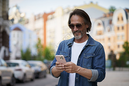 在社交媒体上发短信或发帖 照片是成熟英俊的男人 在户外穿着牛仔衬衫和太阳镜 带着智能手机 英俊的中年男子 头发花白 旅行时拍照图片