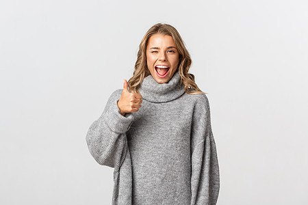 穿着灰色毛衣的有吸引力的快乐妇女肖像 鼓励你 在批准和眨眼中展示拇指 推荐好产品 如并商定白背景图片