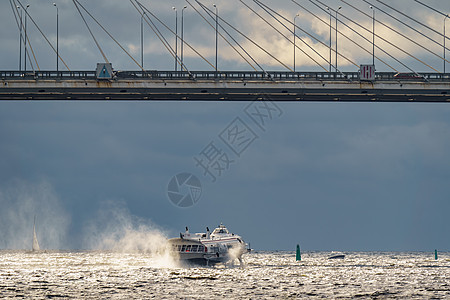 在阳光明媚的天气和背景中的乌云中 一艘高速客乘水流油船在一条有线桥下通过港口日落渡船球拍游客旅行海浪运输巡航旅游图片