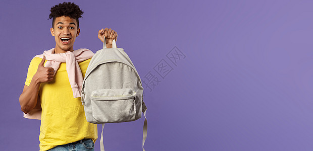 快乐的年轻英俊男学生的画像 男人推荐背包 背着背包 竖起大拇指 为大学学期买了新设备 紫色背景校园教育理发笔记本招聘成人家庭作业图片