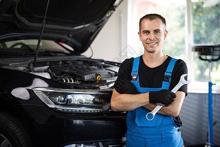 身穿蓝色工作服的年轻白人男子拿着扳手 微笑着看着镜头 宽敞修理店的男性汽车修理工图片
