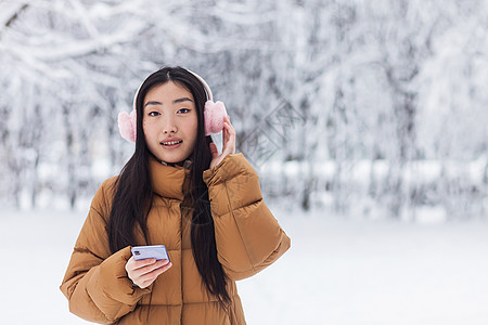美丽的日本青少年在冬季公园散步时使用电话 并用可爱的粉红耳机听音乐和感动的粉红色耳机女士雪花耳朵学生音乐技术手机微笑歌曲粉色图片