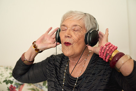 这位奶奶对 Kanye 情有独钟 一位快乐的高级女士在家里的休息室里用耳机听音乐背景