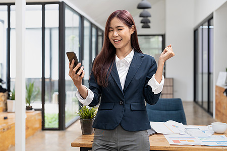 兴奋的亚洲女商务人士挥手拳头 在现代办公室工作时在智能手机上得到好消息图片