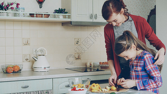 年轻漂亮的妈妈教她可爱的女儿正确切蔬菜 在现代厨房里 小女孩和慈爱的妈妈一起做饭父母幸福孩子木板烹饪胡椒成人家庭母亲训练图片