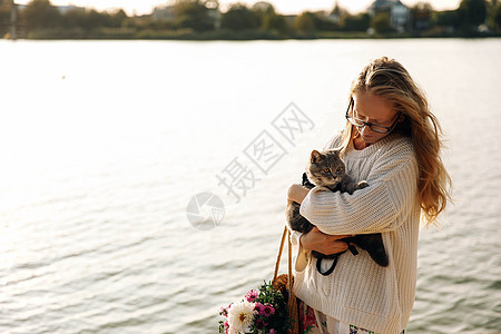 女人金发女金发 在散步时拥抱一只苏格兰直立的小猫家畜动物海滩沙滩猫科动物宠物女性毛衣草包日落图片