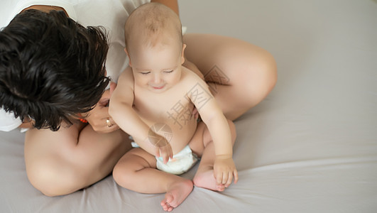 母亲和孩子在一张白色的床上 穿着尿布的妈妈和男婴在卧室里玩耍 父母和小孩在家里放松 一家人在一起玩得很开心婴儿毯子玩具女性护理纺图片