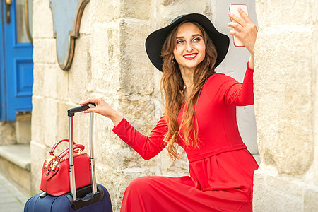 戴着黑帽子的美丽的黑人年轻女性 看着智能手机微笑 坐在户外门口的楼梯上 坐着裙子城市行李相机建筑旅行街道旅游假期航程图片