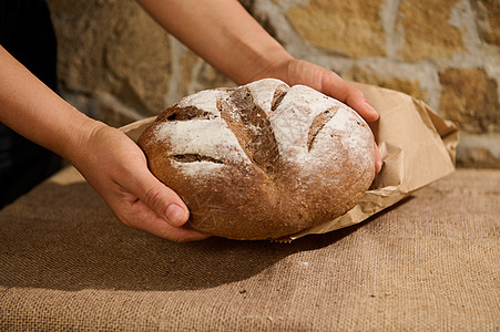 纸袋长棍面包新鲜出炉的自制全麦黑麦面包在家庭主妇 面包师的手中 在一张带麻布桌布的桌子上方背景