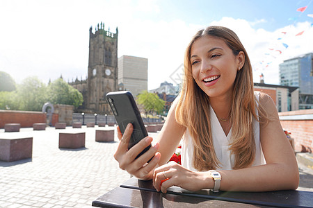 手里拿着手机的微笑女孩坐在户外的桌子旁 背景是城市景观 美丽的年轻女子正在咖啡桌旁的智能手机上观看视频和照片背景图片