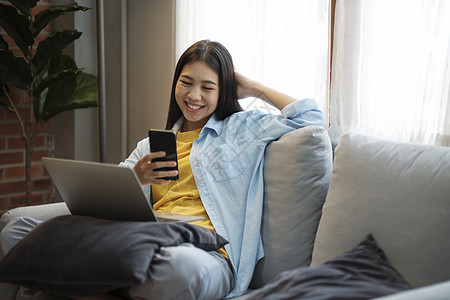 年轻的亚洲女人在家里的沙发上微笑并使用智能手机情感享受说谎阅读房间笔记本活动闲暇客厅冲浪图片