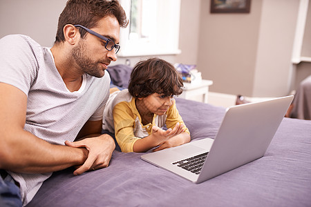 床上电脑玩猜谜游戏 一个父亲和儿子躺在床上时使用笔记本电脑背景
