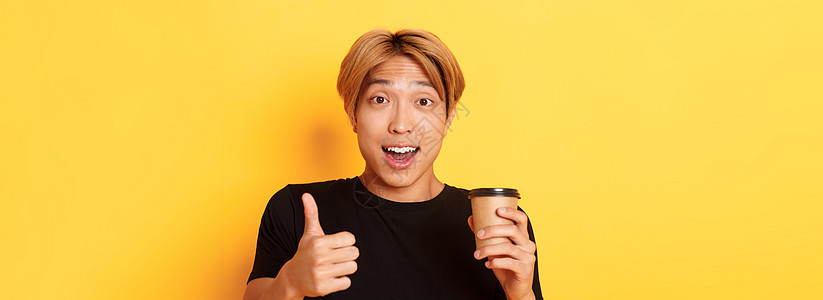 令人惊讶的亚洲英俊帅帅哥推荐咖啡馆 举着咖啡杯和举起拇指来表示赞许 在黄色背景下微笑高兴广告金发学生员工成人快乐情感促销男朋友工图片
