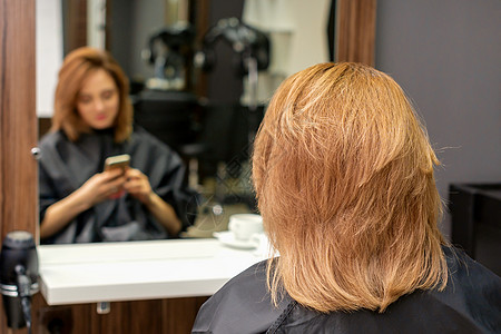 美丽的年轻红发女郎用她的智能手机 在镜子前发短信 等着看理发师的戏呢 笑声女士顾客客户工作室发型师头发女性奢华理发店享受图片
