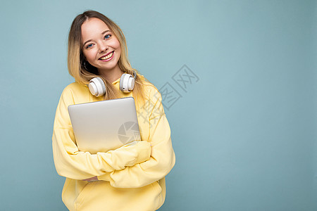 身穿黄色连帽衫的欧洲年轻快乐微笑的女学生手持笔记本 背景为孤立的蓝色背景 有可用空间 教育理念电脑情怀耳机女性成人学生自由职业者图片