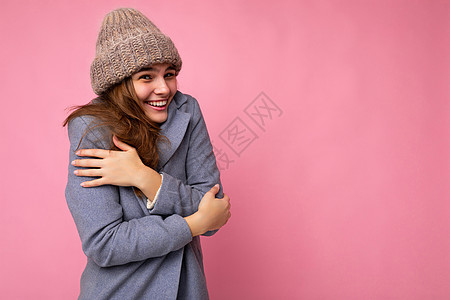 身穿灰色秋大衣和灰顶温暖的帽子 看着相机 感觉寒冷 被粉红色背景墙围着而孤立的年轻黑发美女图片