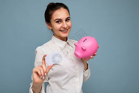 快乐积极微笑的年轻美丽漂亮的黑发女人的画像 带着真诚的情感 穿着休闲的白衬衫 与复制空间隔离在蓝色背景上 拿着粉红色的猪钱盒和塑图片