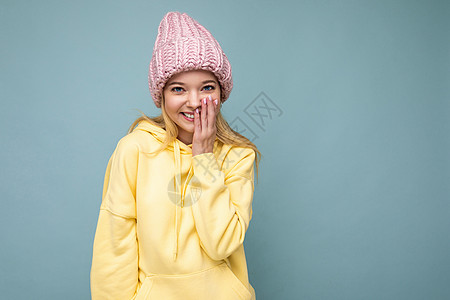 迷人的微笑快乐的金发年轻女性孤立地站在色彩缤纷的背景墙上 穿着日常时尚的服装 看着镜头时表现出面部情绪黄色帽衫可用粉红色蓝色衣服图片