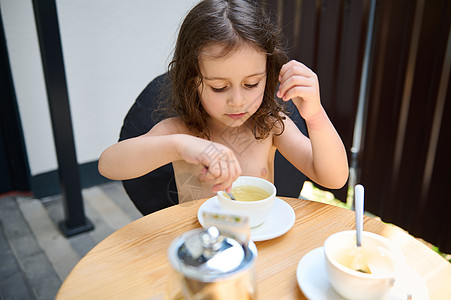 茶杯里小女孩小女孩坐在桌边 喝茶汤匙 在白茶杯里用健康的抗氧化剂草药茶搅拌糖背景