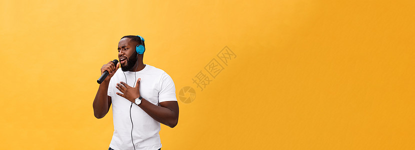 欢快积极 别致 英俊的非洲男子手持麦克风 头戴耳机听音乐唱歌 享受黄色背景中孤立的周末假期的肖像青少年男性女孩女性微笑卷曲播放歌图片