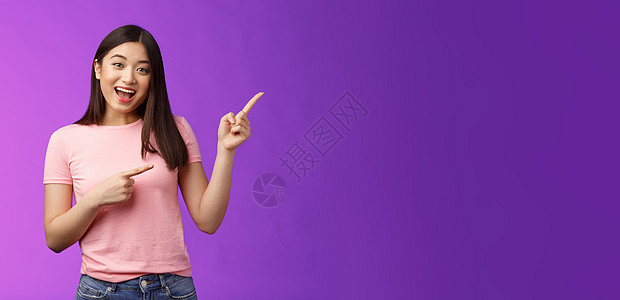 友好而热情的迷人亚洲女孩在进行有趣的谈话 讨论感兴趣的新店 向右指着邀请进来 站在紫色背景微笑的相机上展示黑发情感促销大学旅游学图片