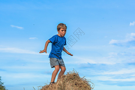 男孩微笑着玩着舞蹈鬼脸炫耀蓝色 T 恤站在干草堆上 晴朗的天空阳光灿烂的日子 平衡训练 快乐童年的概念 户外儿童 接近大自然的清图片
