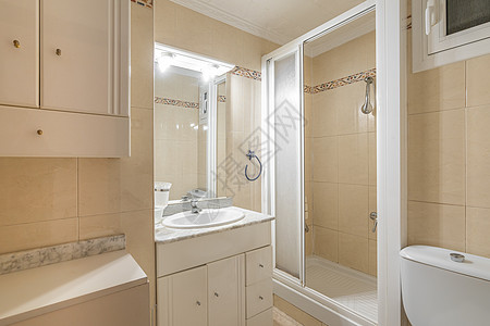 浴室在小镜子上方的灯下显得明亮 墙壁上铺着浅米色瓷砖 带磨砂玻璃推拉门的白色塑料开放式淋浴间 有许多抽屉和白色水槽的梳妆台背景图片
