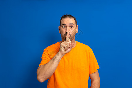 胡子拉蒂诺男子穿着橙色临时T恤 用手指对着嘴唇像要求保持安静一样 隔绝在蓝背景上图片