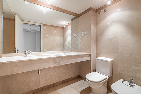 现代宽敞的浴室配有明亮的米色瓷砖 马桶 坐浴盆和长大理石台面上的水槽 穿衣镜映出通往走廊的敞开门 天花板上的灯泡发出明亮的光图片