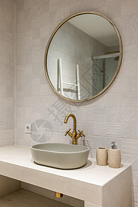 白色大理石台面上整洁时尚的椭圆形水槽 配有优雅的铜色水龙头 水槽上方悬挂着一面圆形的现代金框镜子 可以映出部分淋浴间图片